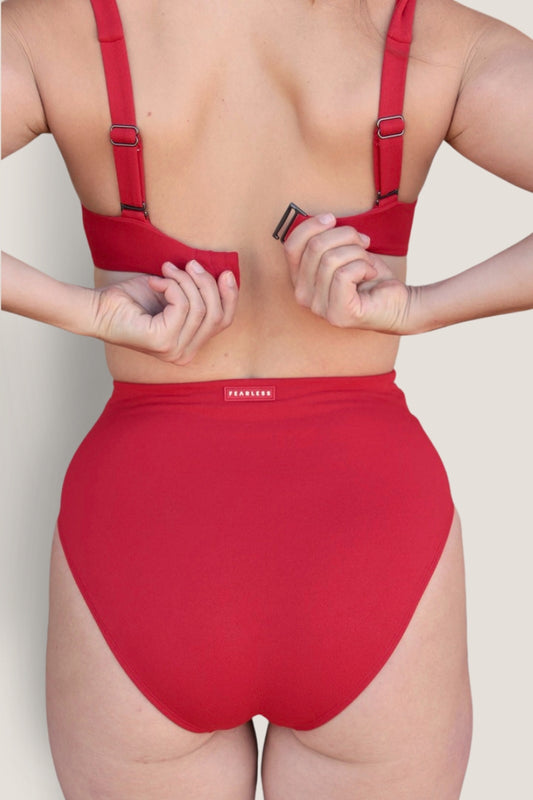 A person seen from the back adjusts the strap of a matching red Icon Bikini Top by Fearless au, paired with a red high-waisted bikini bottom made from recycled fabric.