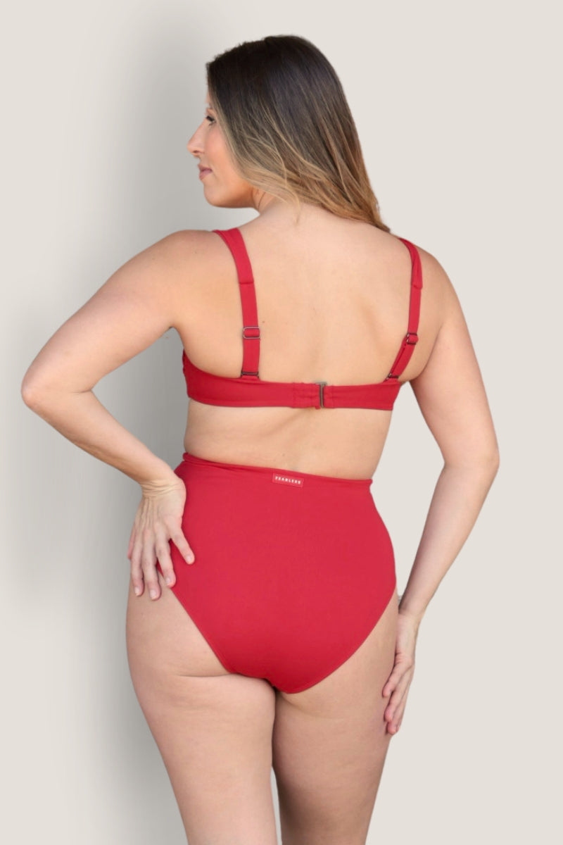 A woman faces away from the camera wearing the Fearless au Icon Bikini Top—a red, high-waisted bandeau bikini with adjustable straps, made from recycled crinkle fabric—set against a plain light gray background.