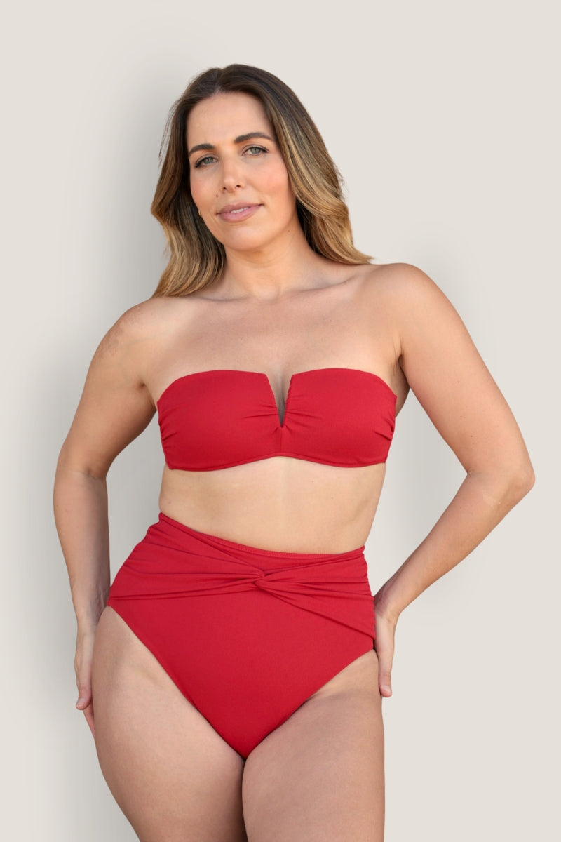 A woman wears the Fearless au Icon Bikini Top, a red strapless bandeau with a V-notch, paired with high-waisted bottoms made from recycled fabric. She stands against a light beige background, looking at the camera with a slight smile.