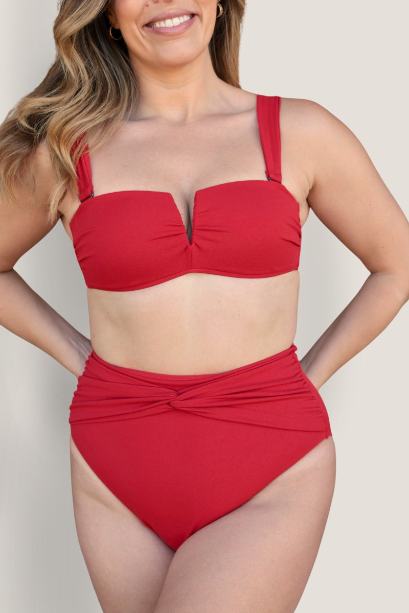 A woman wears the Fearless au Icon Bikini Top—structured with moulded cups and paired with bright red high-waisted bottoms—smiling with her hands behind her back against a plain light background.