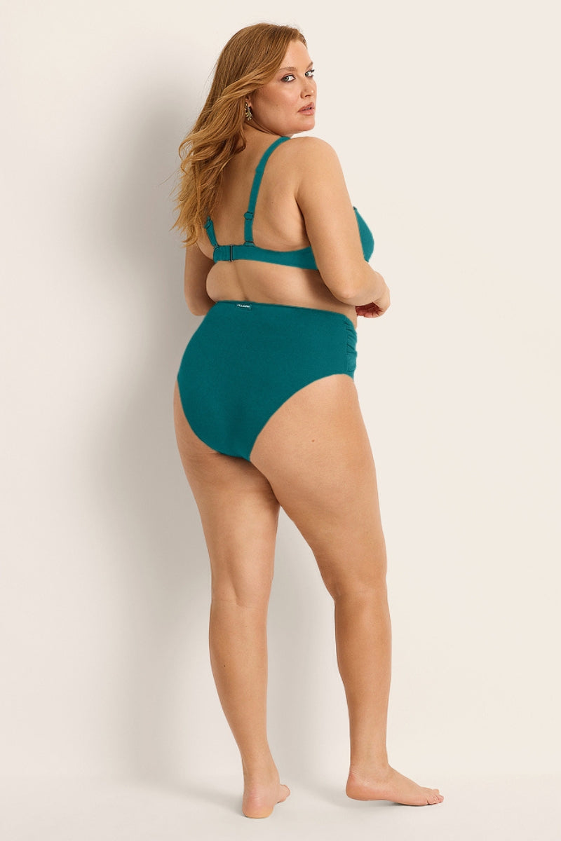 A woman with long, light brown hair poses barefoot in the Fearless au Icon Bikini Top, a teal high-waisted bikini made from recycled fabric, standing sideways and looking over her shoulder against an off-white background.