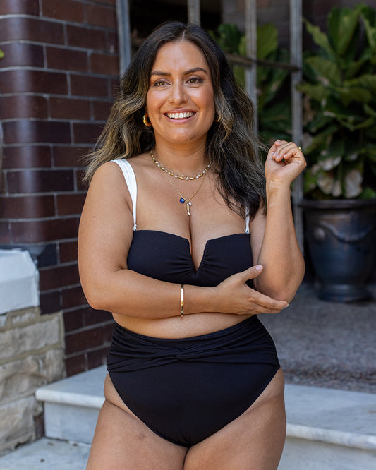 A woman with long, wavy hair smiles outdoors, wearing the Fearless au Icon Bikini Bottom in black and white and layered gold jewelry. She stands in front of a brick wall with a potted plant and stone steps behind her.