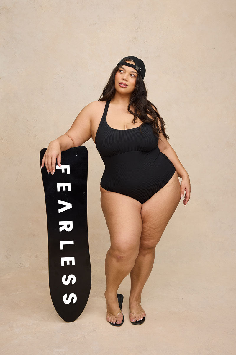 A plus-size woman stands in a studio wearing a black one-piece swimsuit and holding a black surfboard with the word 'FEARLESS' printed vertically in white. She is posed against a warm beige textured backdrop.
