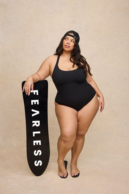 A plus-size woman stands in a studio wearing a black one-piece swimsuit and holding a black surfboard with the word 'FEARLESS' printed vertically in white. She is posed against a warm beige textured backdrop.