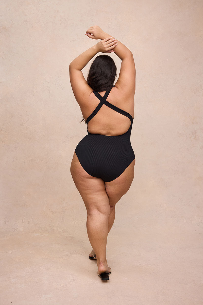 A woman viewed from behind wearing a black one-piece swimsuit with crisscross back straps, standing against a warm beige studio backdrop.