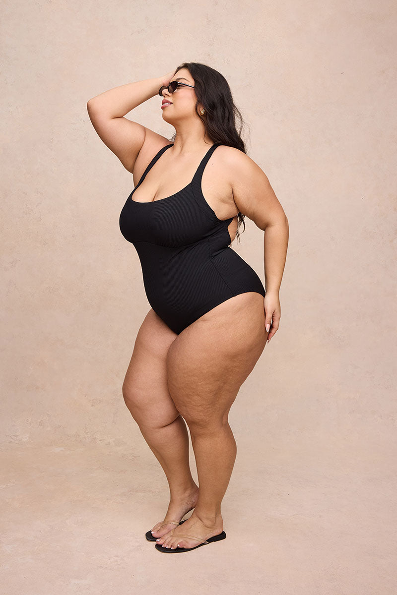 A woman modeling a black one-piece swimsuit against a warm beige textured studio backdrop.