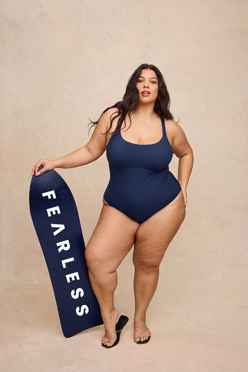 A plus-size woman stands in a studio wearing a navy one-piece swimsuit and holding a navy board with the word 'FEARLESS' in white. The background is a textured beige studio wall.