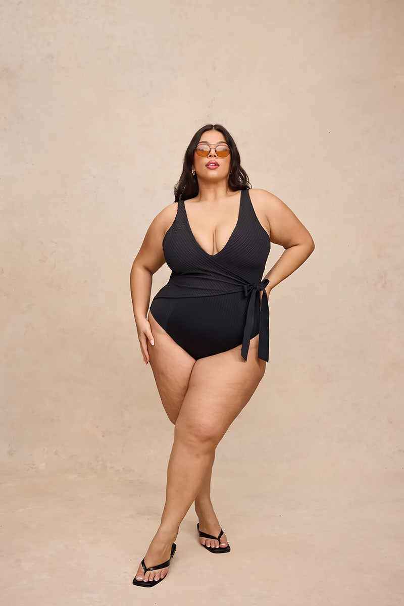 A plus-size woman modeling a black one-piece swimsuit with a deep V-neck, halter-style shoulder straps, and a side tie detail, paired with black sandals against a textured beige studio backdrop.