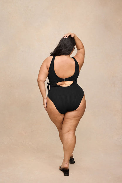A woman is shown from behind wearing a black ribbed one-piece swimsuit with a cutout back, standing against a textured beige studio backdrop.