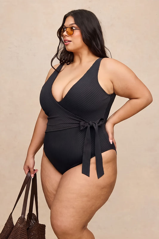 A woman modeling a black ribbed one-piece swimsuit with a wrap-style V-neck, side tie, and side cutout, standing against a plain beige studio background and holding a brown woven tote bag.