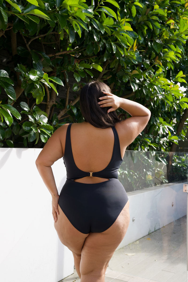 A woman wearing a black one-piece swimsuit with a low open back and side cutout, standing outdoors in front of green foliage and a white wall.