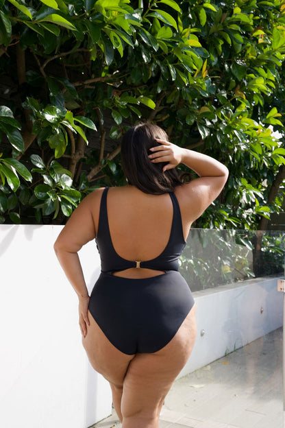 A woman wearing a black one-piece swimsuit with a low open back and side cutout, standing outdoors in front of green foliage and a white wall.