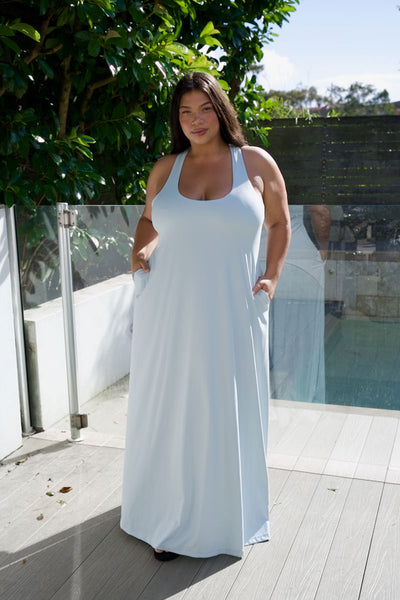 A woman wearing a light blue sleeveless maxi dress with pockets, standing on a wooden deck with a glass railing and green foliage in the background.