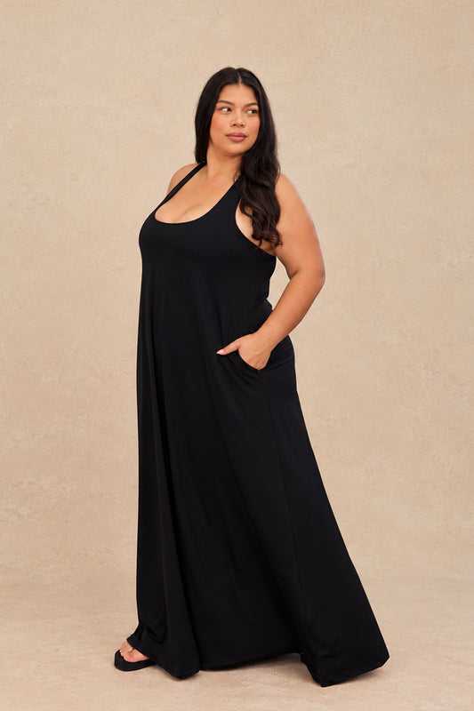 A woman modeling a sleeveless black maxi dress with a scoop neckline and side pockets, standing against a textured beige studio backdrop.