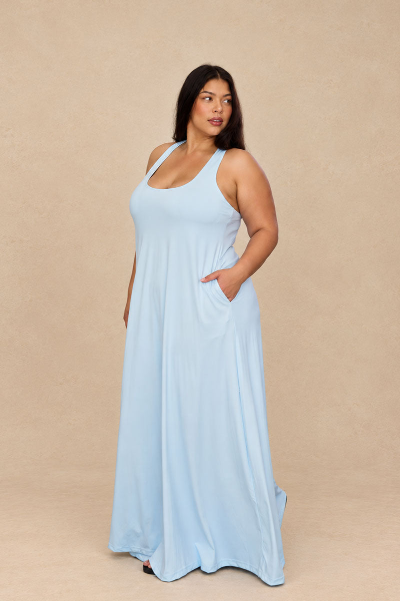 A woman modeling a light blue sleeveless maxi dress with a scoop neckline and side pockets, standing against a solid beige studio backdrop.