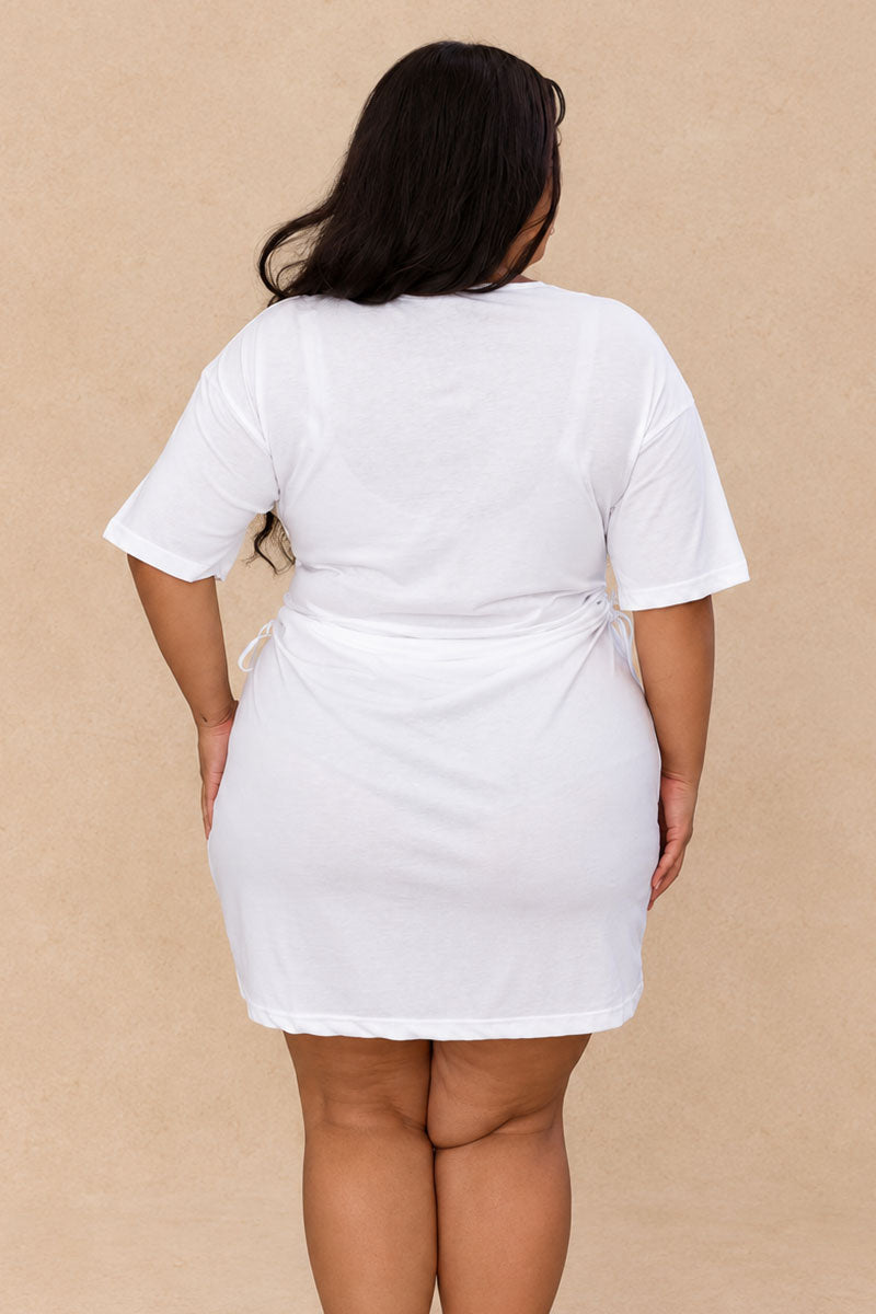 A woman viewed from the back wearing a white short-sleeve mini dress with a tie waist, standing against a plain beige background.