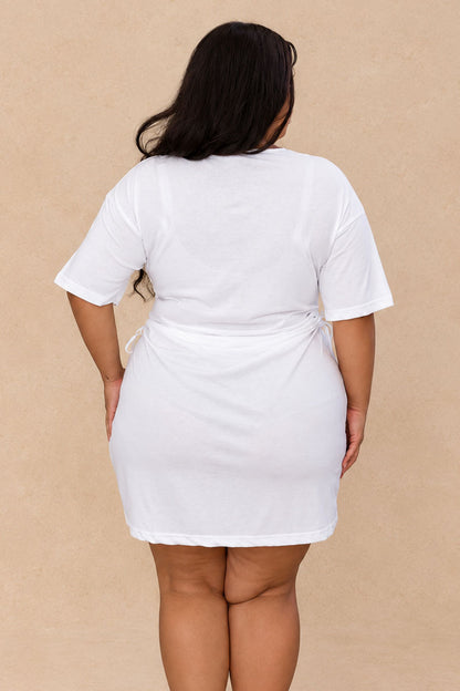 A woman viewed from the back wearing a white short-sleeve mini dress with a tie waist, standing against a plain beige background.