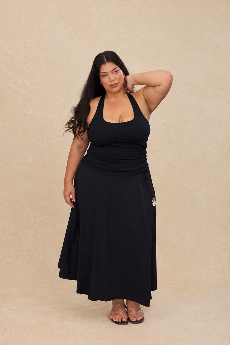 A woman modeling a black tank and skirt with a ruched waist and flared skirt, standing against a textured beige studio backdrop.