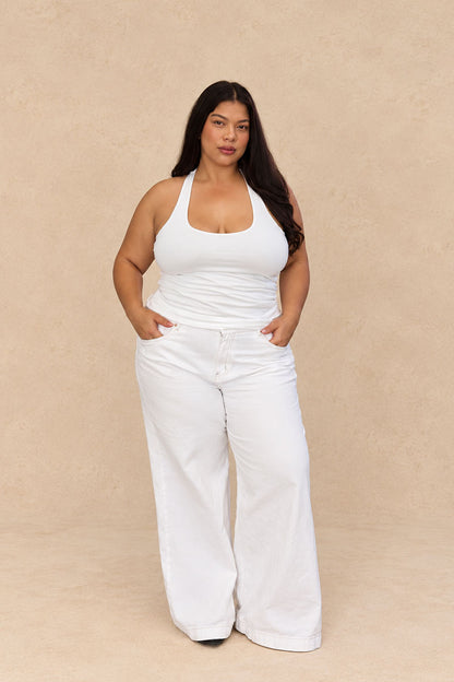 A woman modeling a white sleeveless halter top and matching white wide-leg pants against a plain beige studio backdrop.