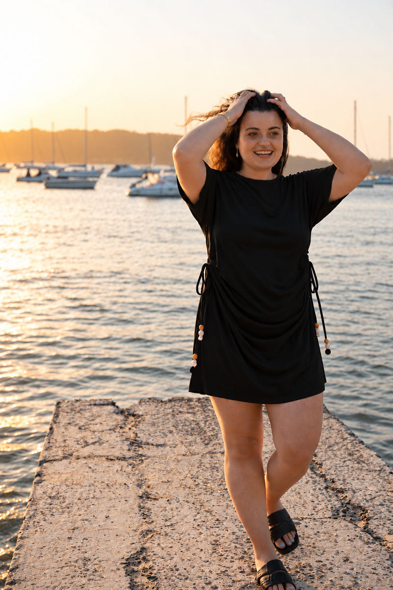 A woman wearing a black short-sleeve mini dress with drawstrings stands on a concrete pier by the water at sunset, with boats visible in the background.