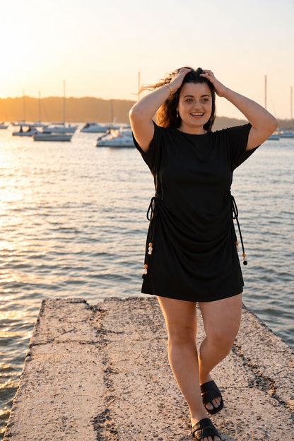 A woman wearing a black short-sleeve mini dress with drawstrings stands on a concrete pier by the water at sunset, with boats visible in the background.