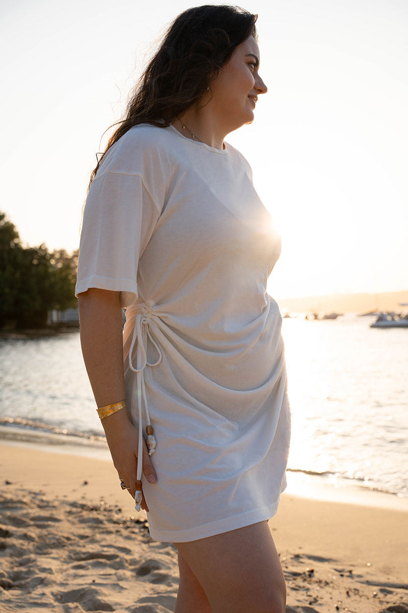 A woman wearing a white short-sleeve t-shirt dress with a side drawstring, standing on a sandy beach at sunset with the ocean in the background.