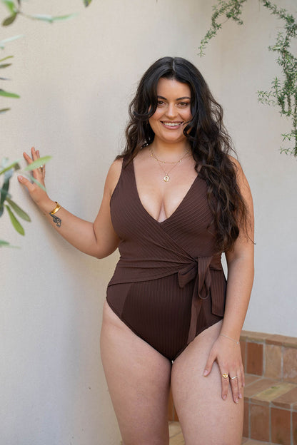 A woman modeling a ribbed brown one-piece swimsuit with a wrap-style V-neck and side tie, standing against a light beige wall with greenery.
