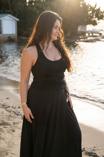 A woman wearing a black sleeveless tank and wrap skirt stands on a sandy beach near the water, with warm sunlight and a blurred outdoor background.