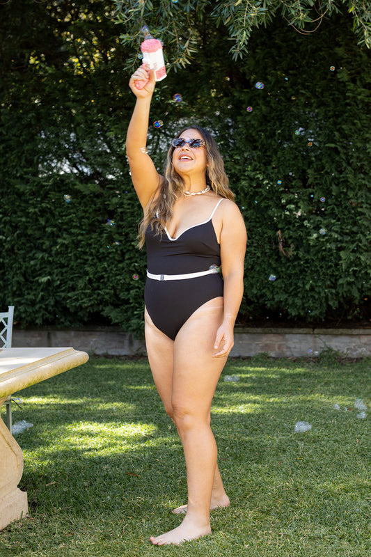 A woman stands barefoot on grass, smiling and holding a pink bubble wand as bubbles float around her. She wears the Fearless au Grace High Rise one-piece swimsuit in Matte Black. Greenery and a stone table are nearby.