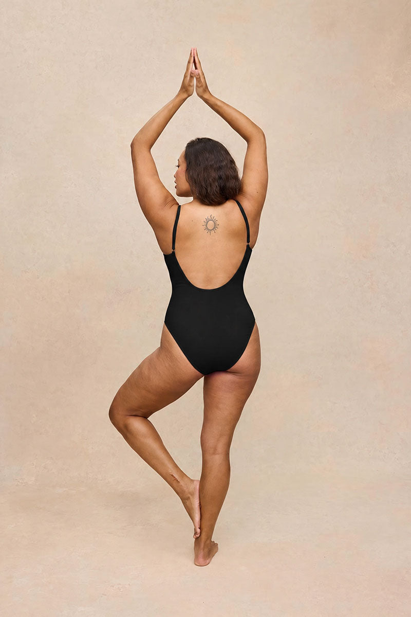 A woman wearing a black one-piece swimsuit or leotard stands in a yoga pose from the back against a warm beige textured studio background.