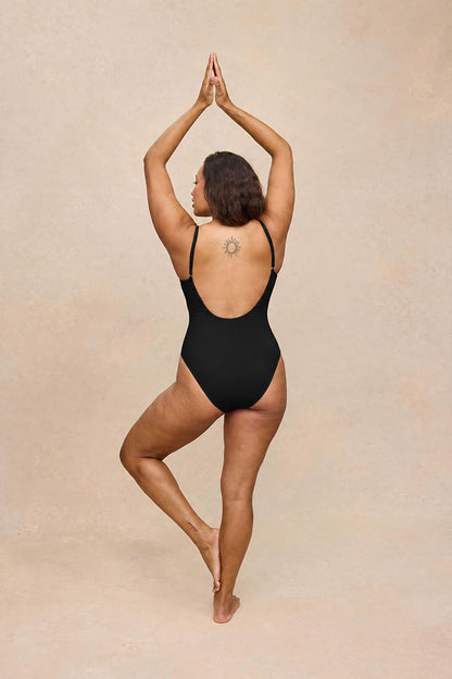 A woman wearing a black one-piece swimsuit or leotard stands in a yoga pose from the back against a warm beige textured studio background.