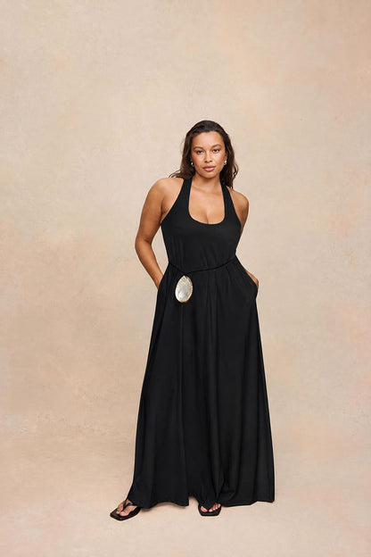 A woman modeling a black halter-neck maxi dress with side pockets and a large silver circular belt accent, standing against a textured beige studio backdrop.