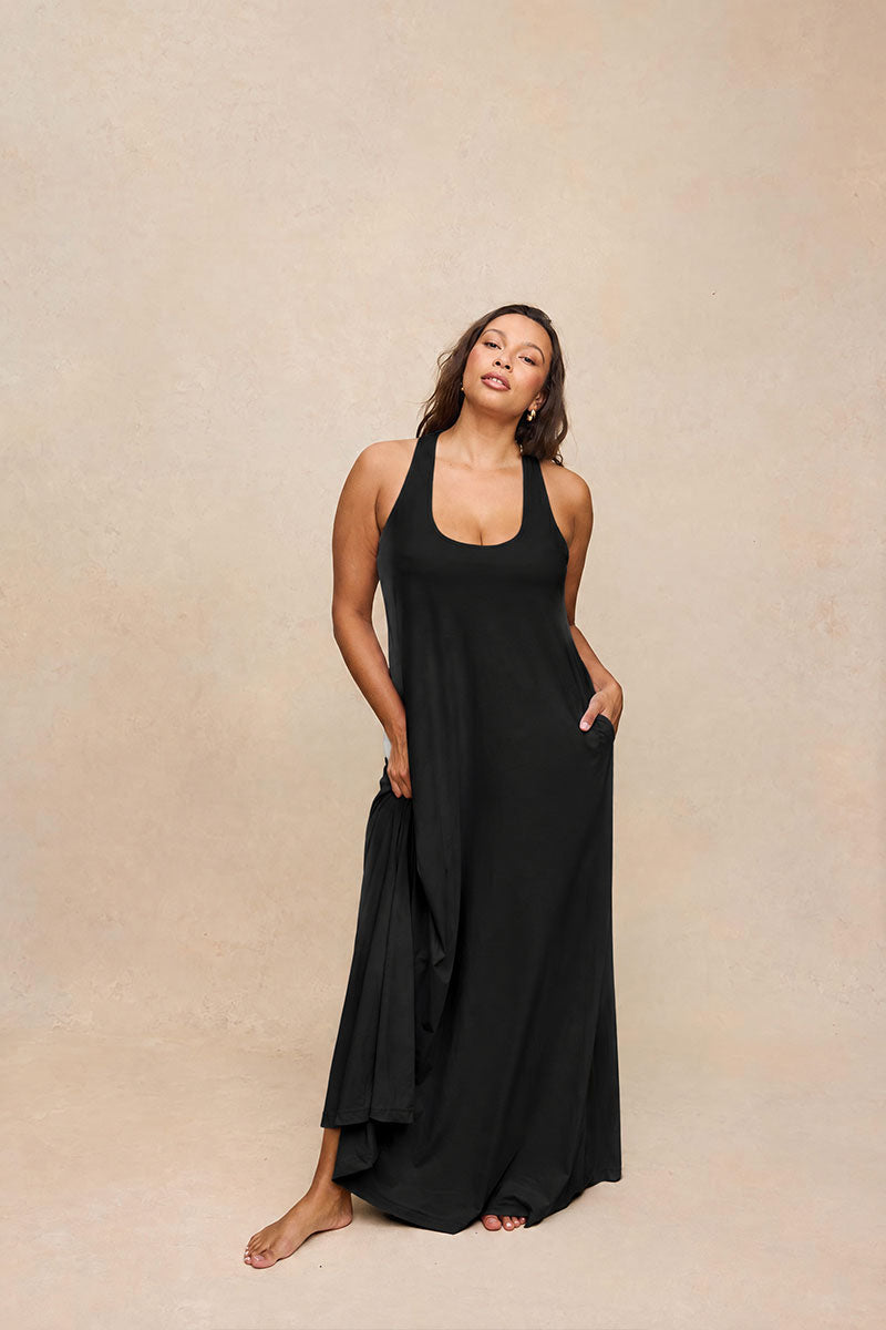 A woman modeling a black halter-neck maxi dress with side pockets, standing against a textured beige studio backdrop.
