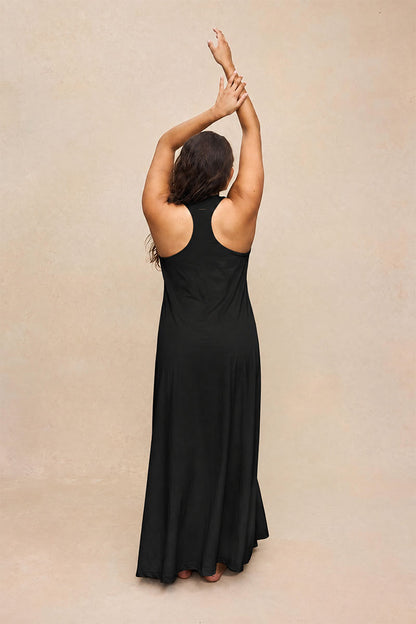A woman wearing a sleeveless black maxi dress, shown from behind with her arms raised overhead, standing against a textured beige studio backdrop.