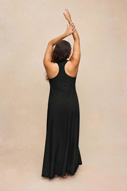 A woman wearing a sleeveless black maxi dress, shown from behind with her arms raised overhead, standing against a textured beige studio backdrop.