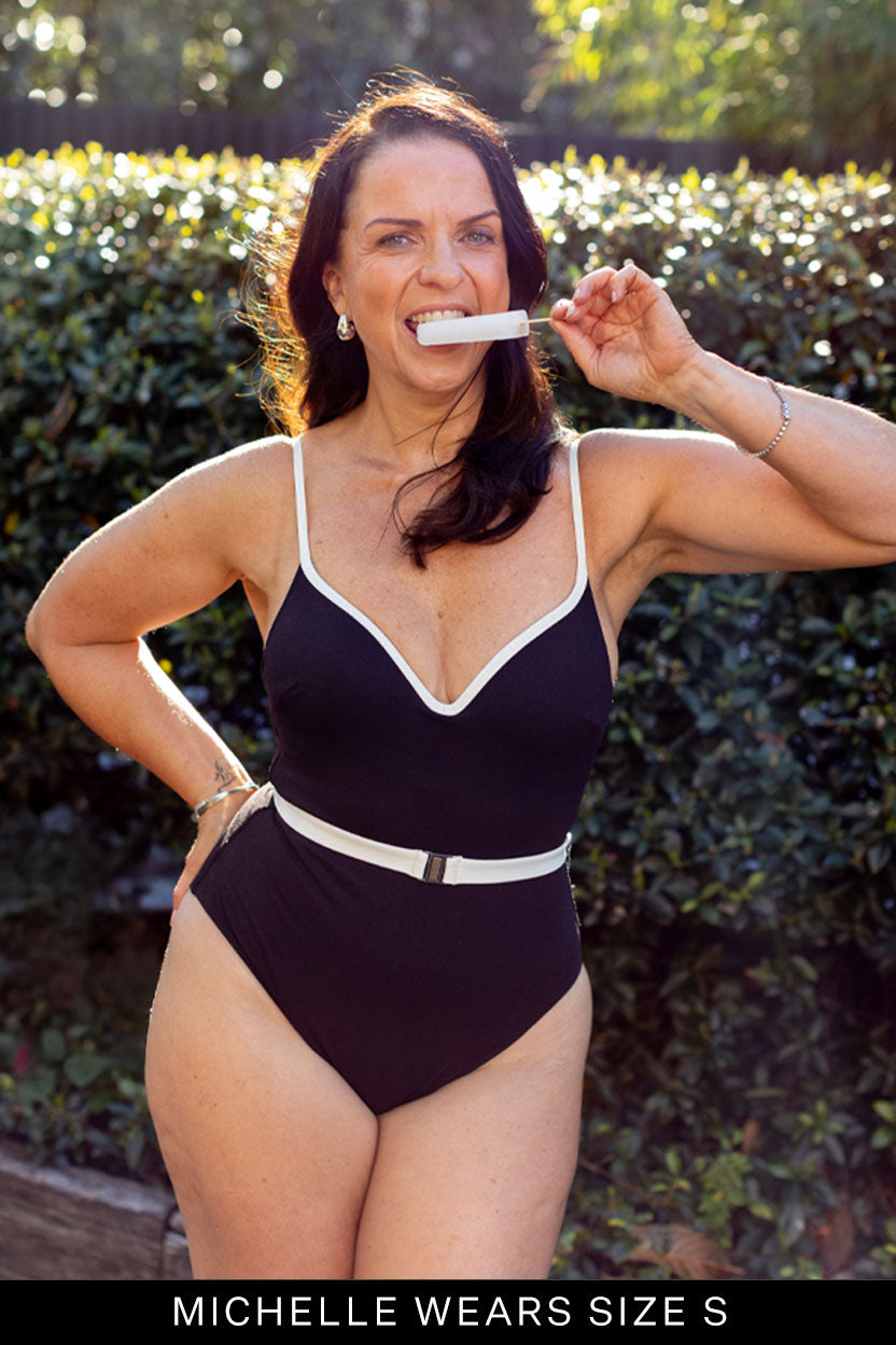 A woman in a Fearless au Grace High Rise One Piece - Standard swimsuit stands outdoors before greenery, smiling confidently with a tube-shaped item in her mouth. Text below reads: MICHELLE WEARS SIZE S.