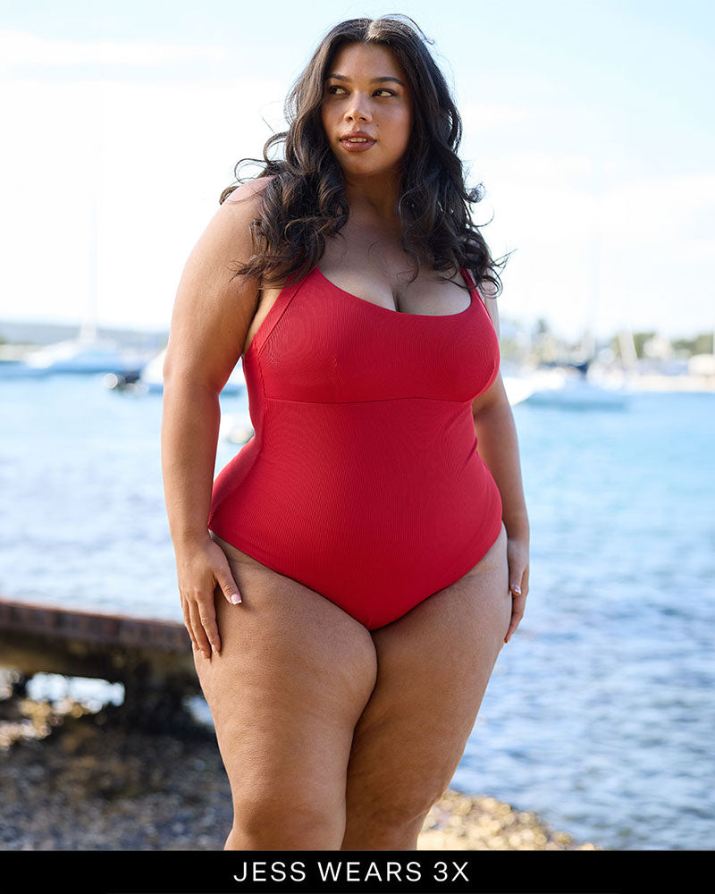 Woman in a Fearless red supportive bust and one piece swimsuit standing by a waterfront, sunlit background