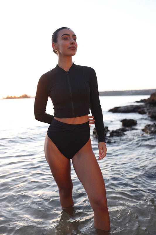 Person wearing a black swimsuit standing in water near a rocky shore.