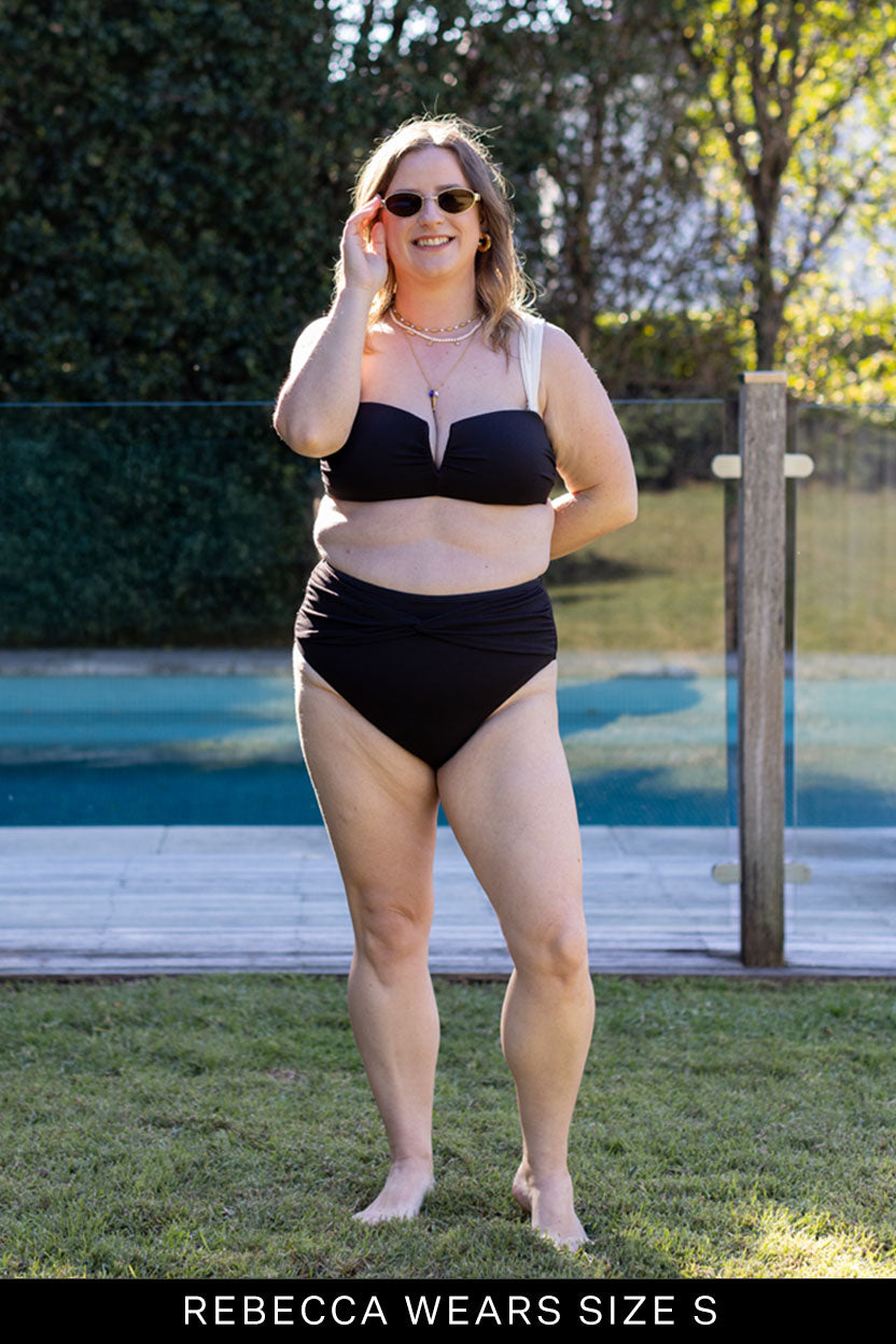 A woman stands on grass near a pool, smiling at the camera in Fearless au's Icon Bikini Top (black, strapless, moulded cups) and sunglasses. Text at the bottom reads, “REBECCA WEARS SIZE S.”.