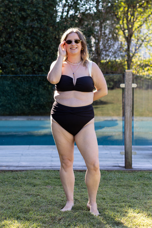 A woman wearing the Fearless au Icon Bikini Top and black sunglasses stands barefoot on grass by a pool, smiling with one hand touching her face on a sunny day.