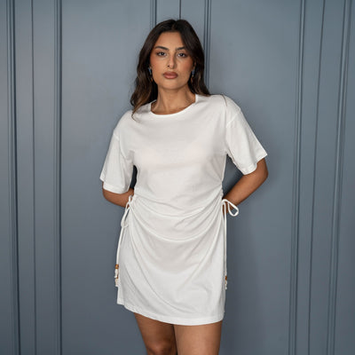 A woman modeling a white short-sleeve wrap-style mini dress with side drawstrings, standing against a gray paneled wall.