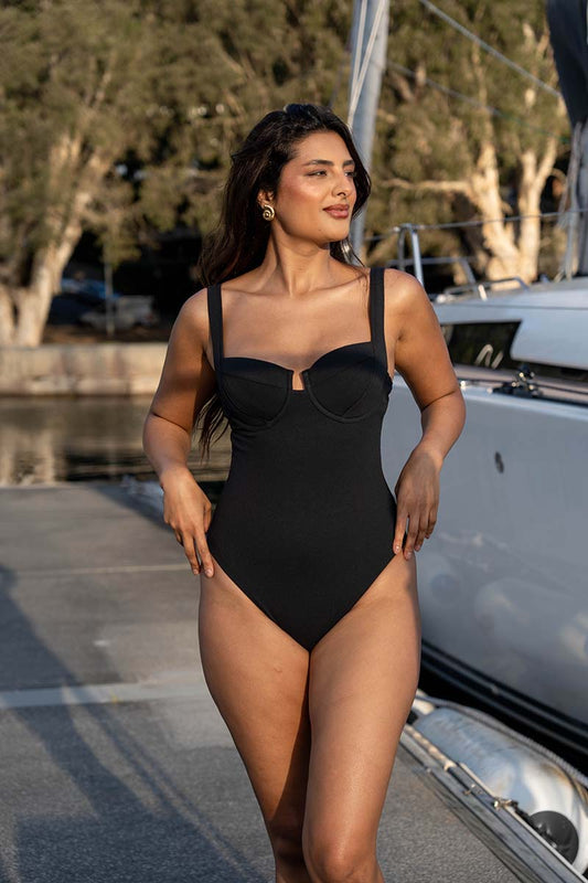 A woman modeling a black one-piece swimsuit on a boat deck next to a white sailboat, with trees and water in the background.