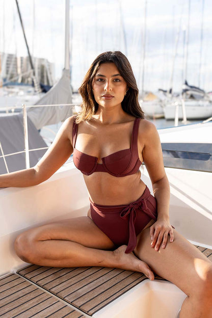 A woman wearing a two-piece maroon bikini sits cross-legged on the white deck of a yacht, with a marina background of boats and water.