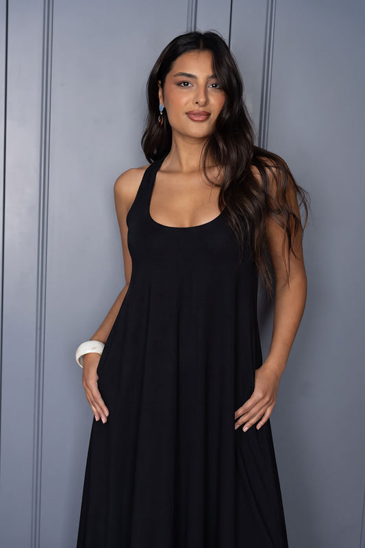 A woman modeling a sleeveless black maxi dress with a scoop neckline, standing against a gray paneled wall.