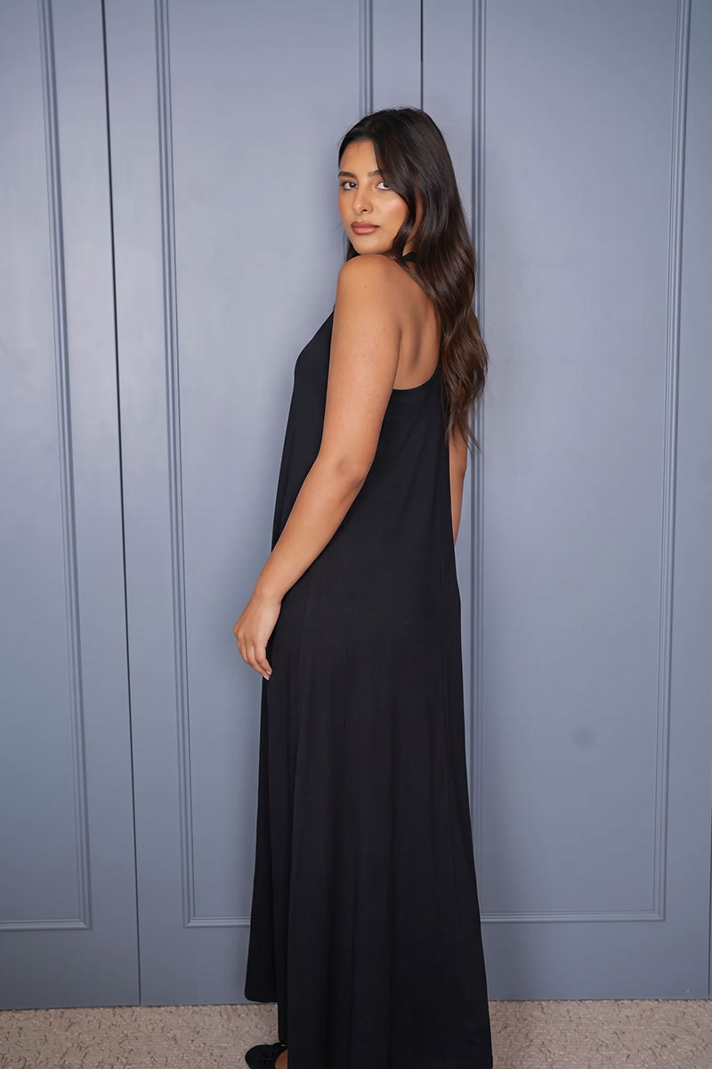 A woman wearing a sleeveless black maxi dress, photographed from the back against a blue-gray paneled wall.
