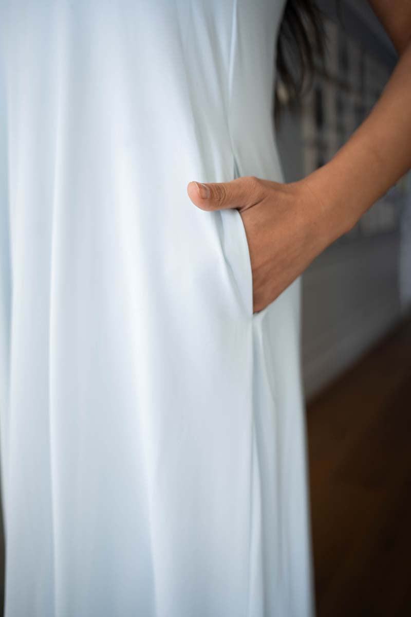 A close-up of a white flowing garment with a side pocket, shown on a person whose hand is reaching into the pocket. The background is softly blurred with a hint of a staircase and wooden floor.