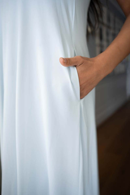 A close-up of a white flowing garment with a side pocket, shown on a person whose hand is reaching into the pocket. The background is softly blurred with a hint of a staircase and wooden floor.