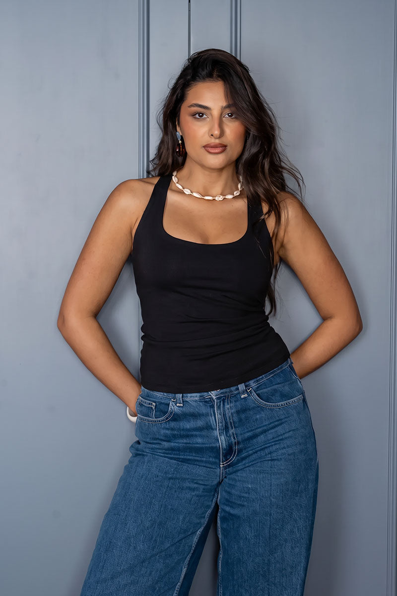 A woman wearing a black ribbed tank top and blue jeans, posing against a gray paneled wall.