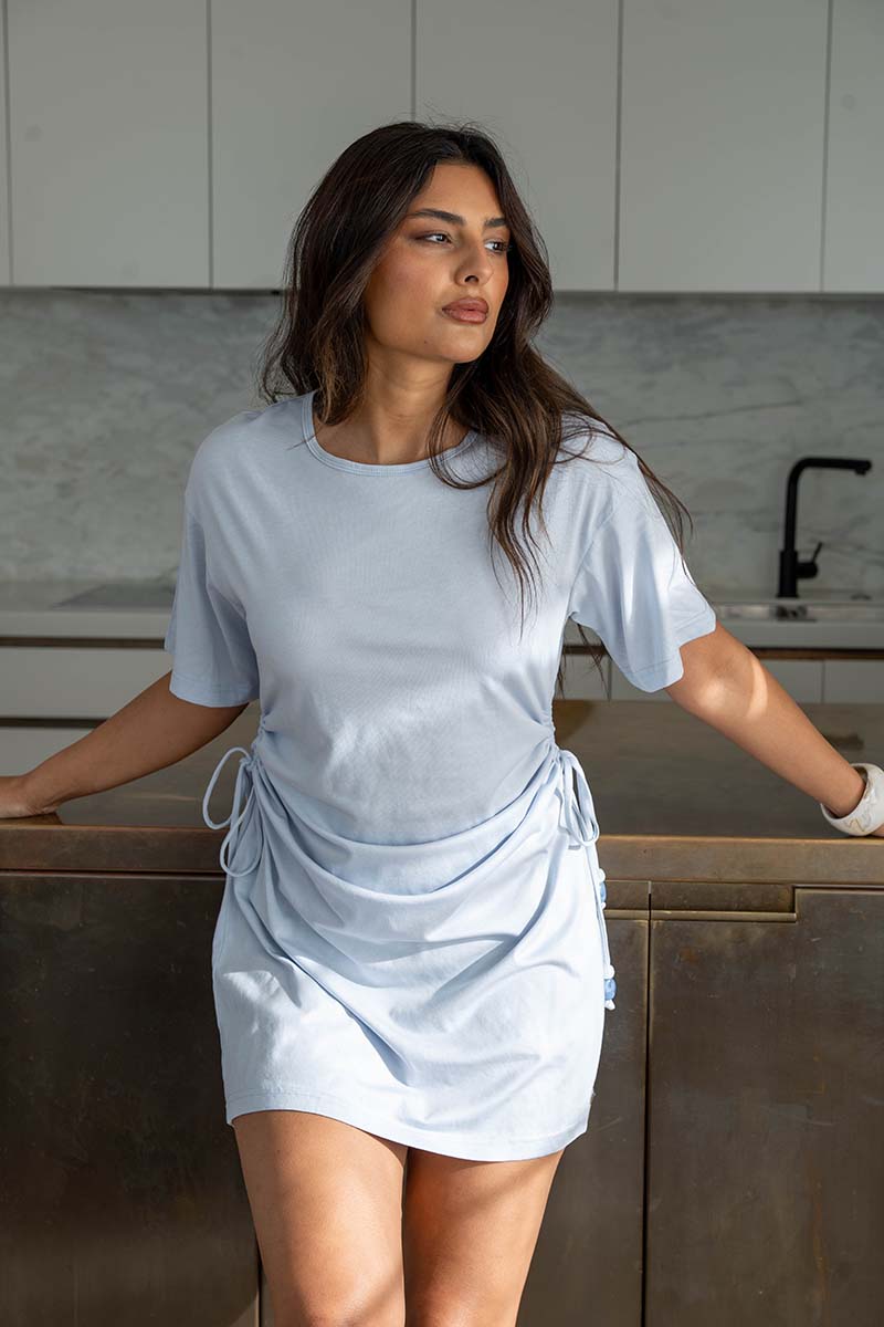 A woman wearing a light blue t-shirt dress with a wrap-style skirt and side drawstrings, posing in a modern kitchen setting.