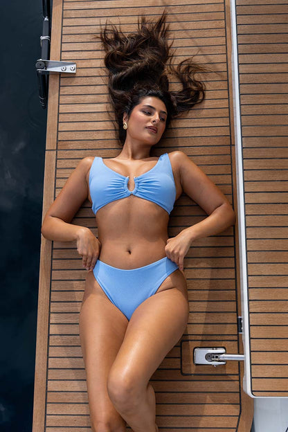 A woman wearing a light blue two-piece bikini swimsuit lies on her back on a wooden boat deck next to the water.